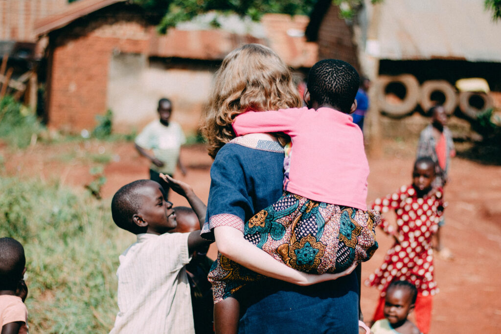 Caucasian woman volunteer in blue dress carrying little girl in pink shirt on her back through the village.