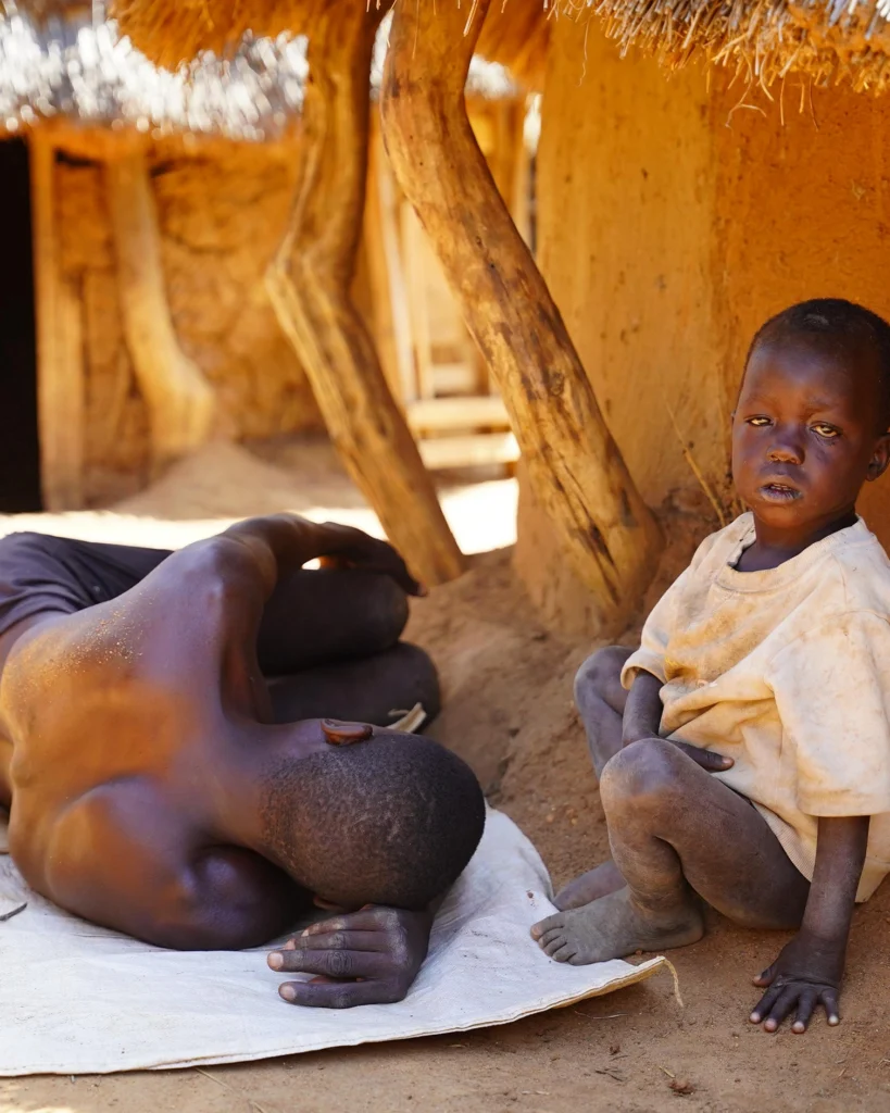 Young child sitting beside an adult outside their home in rural Uganda.